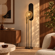 Lampadaire Totem en métal noir et doré H146 cm, allumé dans un salon aux murs en lambris bois à côté d'un fauteuil blanc