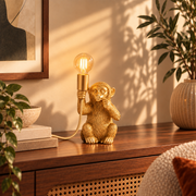 Lampe à poser Monkey en plastique doré, allumée sur un buffet en bois et cannage entouré de plantes